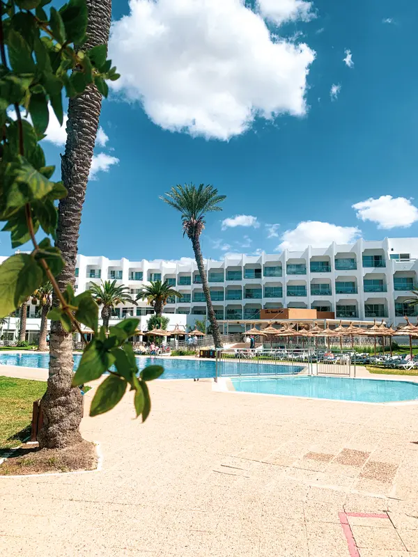 Outdoor pool and white hotel facade with palm trees under blue sky — Aziz Stiti hotel photography